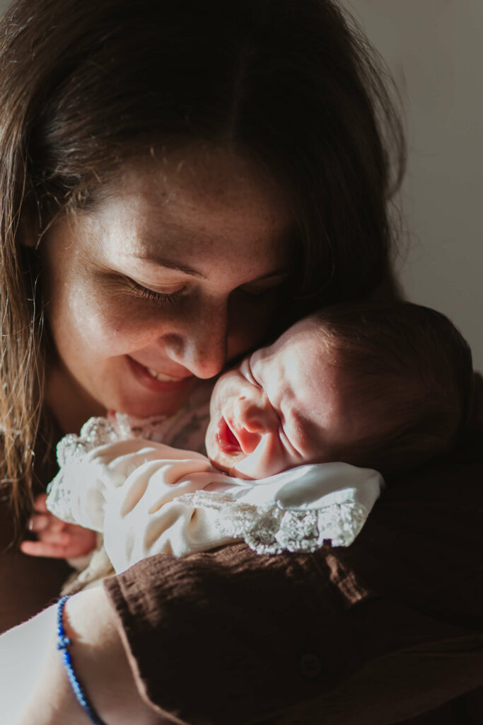 Fotografisanje beba kod kuće – mama drži svoju bebu pored prozora uz prirodno svjetlo.