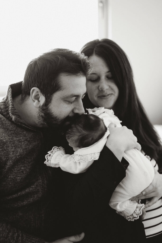 Fotografisanje beba kod kuće – mama i tata drže svoju bebu pored prozora uz prirodno svjetlo